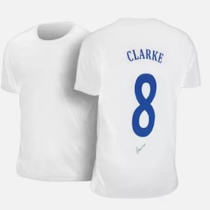 Może przedstawiać: Biała koszulka z nadrukiem imienia "CLARKE" i numeru "8" na plecach w kolorze niebieskim. Koszulka jest podpisana niebieskim atramentem.
