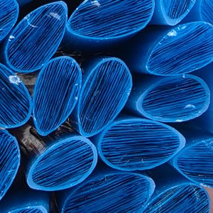 May include: A bundle of numerous individual blue plastic rods, each featuring an oval cross-section. The ends of the rods display a textured, fibrous appearance with fine parallel lines visible across the surface of each cut end.