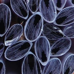 May include: A bundle of numerous thin, cylindrical glass rods with a translucent lavender exterior and a dark, opaque black core. Each rod displays a distinct fibrous, streaked pattern within the lavender glass surrounding the solid black center.