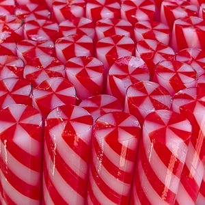 May include: A collection of cylindrical hard sweets arranged in a dense cluster. Each sweet features a repeating pattern of vibrant red and white diagonal stripes that converge into a star-like design on the flat top surface of the cylinder.