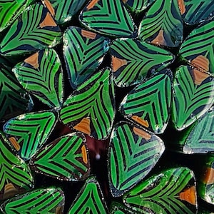 Può includere: Primo piano di una collezione di perline di vetro verdi e nere. Ogni perlina ha un design a forma di foglia con strisce verdi e nere e un accento marrone. Le perline sono disposte vicine, creando una superficie strutturata.