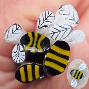 May include: Close-up of handmade glass beads with a bee theme. The beads feature yellow and black stripes, and white leaf designs. The glass beads are held in a hand, showcasing the intricate details and craftsmanship.