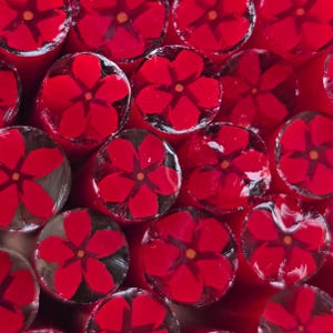 May include: Close-up of red floral candy. Each piece features a vibrant red flower design with yellow centers, encased in a clear, glossy coating. The candies are arranged closely together, creating a repeating pattern against a red background.