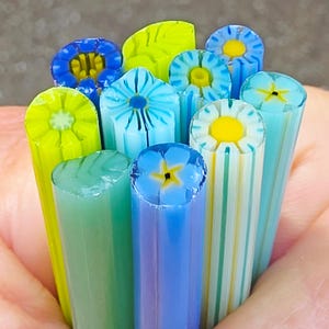 May include: A collection of cylindrical glass canes featuring intricate millefiori patterns in shades of lime green, sky blue, royal blue, and yellow. Each rod displays a unique floral or geometric cross-section design with vibrant, translucent colors.