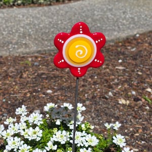 May include: A red flower garden stake with a yellow and white center. The flower has six petals with white dots and is mounted on a thin metal rod. The stake is placed in a garden bed with white flowers and brown mulch.
