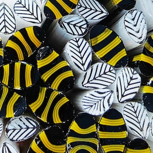 May include: A collection of cylindrical polymer clay canes arranged together. Some canes feature a repeating black and yellow horizontal striped pattern, while others display a white and black leaf-like herringbone design on their circular ends.