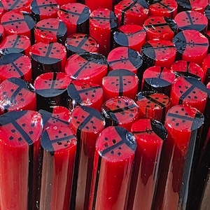 May include: A collection of cylindrical red glass rods arranged vertically. Each rod features a distinct black ladybug pattern on its circular top surface, characterized by a central dividing line and small black dots on a vibrant red background.