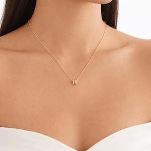 May include: A gold-coloured necklace featuring a fine chain and a small pendant shaped like the letter "H". The necklace is worn around the neck, with the pendant centrally placed. The background is blurred.