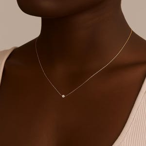 Puede incluir: Delicado collar de oro con un pequeño colgante redondo de piedra blanca. El collar se lleva cerca del cuello, creando un look minimalista y elegante. La cadena es fina y parece ser de oro.