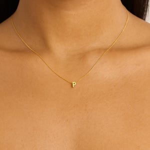 May include: A delicate gold necklace featuring a small letter "P" pendant. The necklace is worn around the neck, with the pendant visible. The chain is fine and elegant, creating a minimalist aesthetic.