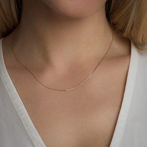 Puede incluir: Un delicado collar de cadena de oro, usado por una modelo, cuelga sobre un fondo blanco.