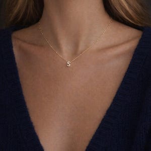 May include: A gold necklace with a small, sparkling letter "S" pendant. The necklace is worn against a dark blue top. The fine chain and pendant are the focal point. The necklace is a subtle, elegant piece.