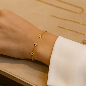May include: A delicate gold bracelet with clover and square charms is displayed on a wrist. Other gold jewelry, including necklaces and earrings, are visible in the background. The jewelry is set against a neutral background.