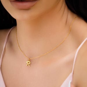 May include: A gold-coloured necklace featuring a fine chain and a small pendant shaped like the letter "R". The pendant hangs centrally, offering a simple, elegant appearance. The necklace is set against a pale pink backdrop.