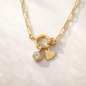 Puede incluir: Collar dorado con un cierre grande y dos colgantes. Un colgante es un cristal redondo y transparente, y el otro es un corazón con la letra "Z" grabada. La cadena tiene un diseño de eslabones rectangulares.