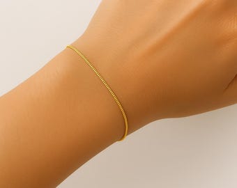 May include: A delicate gold chain bracelet is worn on a wrist. The bracelet is a fine, simple design, with a subtle shine. The background is a neutral, off-white color, highlighting the jewelry.