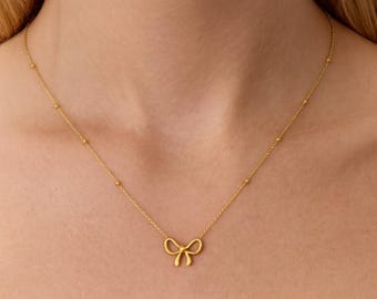 May include: A delicate gold-colored necklace featuring a bow pendant. The necklace has a fine chain with small, evenly spaced beads. The bow pendant is centered and adds a touch of elegance to the jewelry.