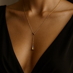 May include: A gold pendant necklace featuring a teardrop charm. The necklace has a fine chain that hangs from a small bar, with a teardrop-shaped pendant. The jewelry is set against a dark background.