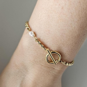 Pode incluir: Uma pulseira dourada com fecho de alternância e um pequeno detalhe de pérola. A pulseira, com um design semelhante a uma corrente, é usada no pulso. O fecho tem um design circular com uma barra. Cerca de 18 cm de comprimento.
