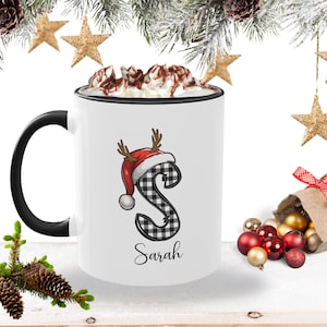 May include: White ceramic mug with a black handle and rim, filled with whipped cream and chocolate drizzle. The mug features a plaid letter "S" wearing a Santa hat with antlers, and the name "Sarah" below.