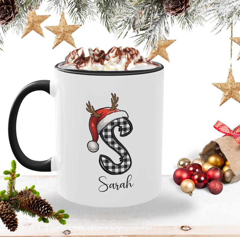May include: White ceramic mug with a black handle and rim, filled with whipped cream and chocolate drizzle. The mug features a plaid letter "S" wearing a Santa hat with antlers, and the name "Sarah" below.