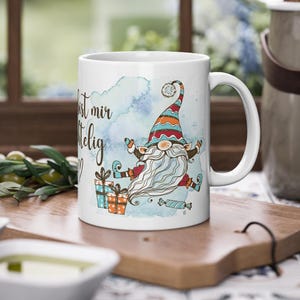Puede incluir: Taza de cer&aacute;mica blanca con un dise&ntilde;o de gnomo caprichoso con una larga barba blanca, un sombrero colorido y regalos. La taza tiene el texto "Du bist mir wichtig" en una fuente de escritura, sobre un fondo azul acuarela.