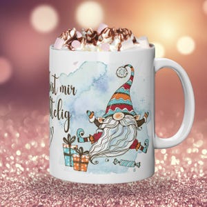 Puede incluir: Taza de cer&aacute;mica blanca con un dise&ntilde;o de gnomo caprichoso, una larga barba blanca, un sombrero colorido y cajas de regalo. La taza est&aacute; llena de crema batida, malvaviscos y salsa de chocolate. La taza tiene el texto "ist mir heilig".