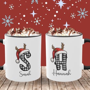 May include: Two white ceramic mugs with black handles and rims, each topped with whipped cream and chocolate shavings. The mugs feature a letter in a black and white plaid pattern, a red Santa hat with antlers, and a name below. The background is red with snowflakes.