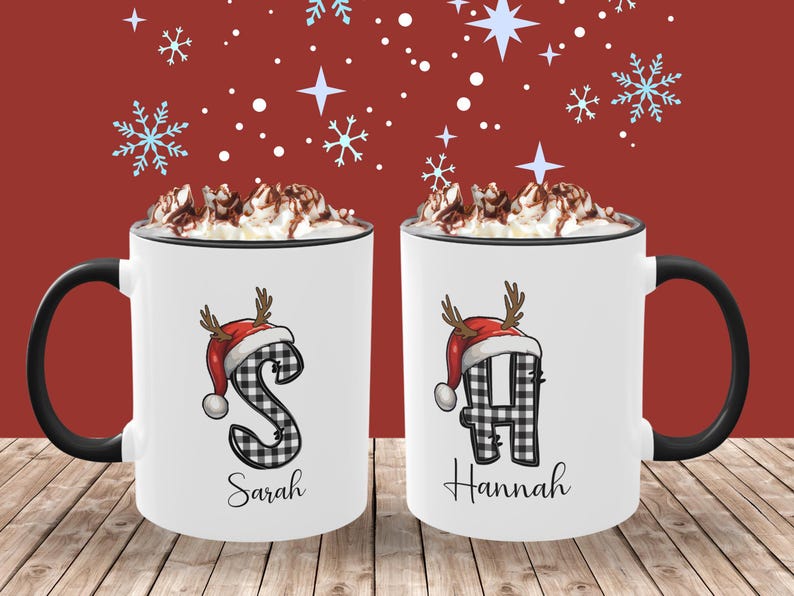 May include: Two white ceramic mugs with black handles and rims, each topped with whipped cream and chocolate shavings. The mugs feature a letter in a black and white plaid pattern, a red Santa hat with antlers, and a name below. The background is red with snowflakes.