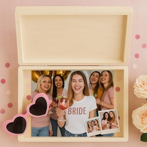 May include: A wooden box with a hinged lid, open to reveal a photograph of a bride and her friends. Pink heart-shaped sunglasses rest on the box. The bride wears a white t-shirt with the word "BRIDE".