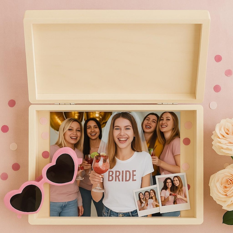 May include: A wooden box with a hinged lid, open to reveal a photograph of a bride and her friends. Pink heart-shaped sunglasses rest on the box. The bride wears a white t-shirt with the word "BRIDE".