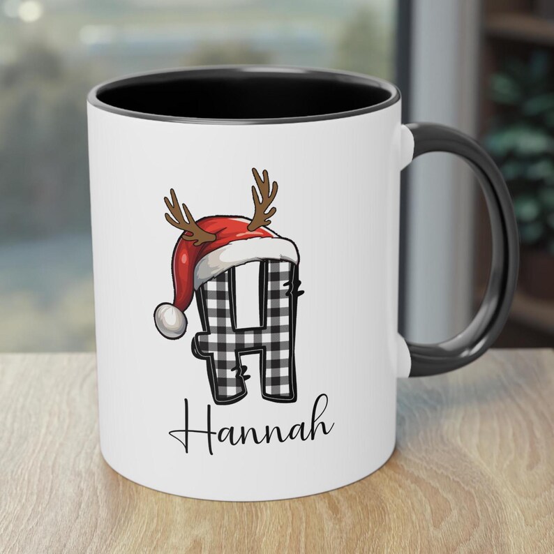 May include: White ceramic mug with a black interior and handle. The mug features a black and white plaid letter "H" wearing a red Santa hat with antlers. The name "Hannah" is written in a cursive font below the letter.