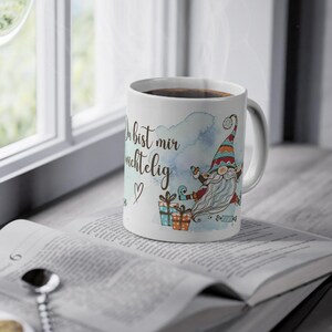 Puede incluir: Taza de cer&aacute;mica blanca con l&iacute;quido oscuro, adornada con una ilustraci&oacute;n de gnomo y texto en alem&aacute;n. El gnomo tiene una larga barba blanca y un sombrero colorido. La taza est&aacute; sobre un libro abierto.