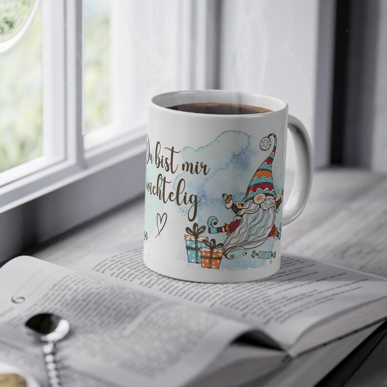 Puede incluir: Taza de cer&aacute;mica blanca con l&iacute;quido oscuro, adornada con una ilustraci&oacute;n de gnomo y texto en alem&aacute;n. El gnomo tiene una larga barba blanca y un sombrero colorido. La taza est&aacute; sobre un libro abierto.