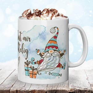 Puede incluir: Taza de cer&aacute;mica blanca con una ilustraci&oacute;n caprichosa de gnomo, texto y cobertura de crema batida con salsa de chocolate. El gnomo lleva un sombrero colorido y sostiene regalos. La taza est&aacute; sobre una superficie de madera.