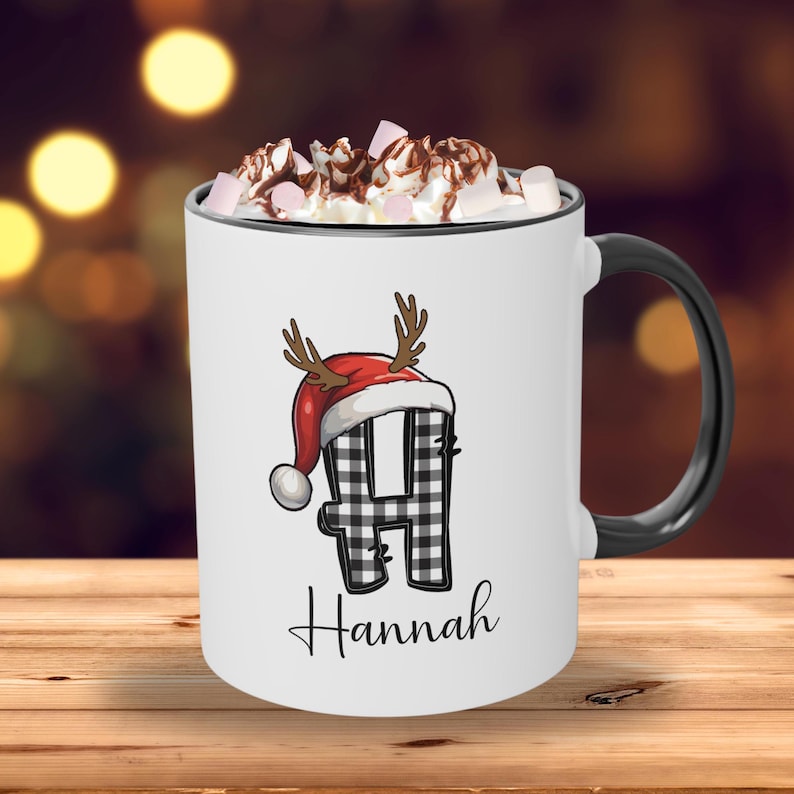 May include: White ceramic mug with a black handle and rim, filled with whipped cream and marshmallows. The mug features a black and white plaid letter "H" wearing a Santa hat with antlers, and the name "Hannah" below.