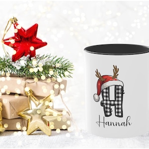 May include: White ceramic mug with a black interior and handle, featuring a plaid letter "H" wearing a Santa hat and antlers. The name "Hannah" is written below. Christmas decorations, including a red star ornament, are in the background.
