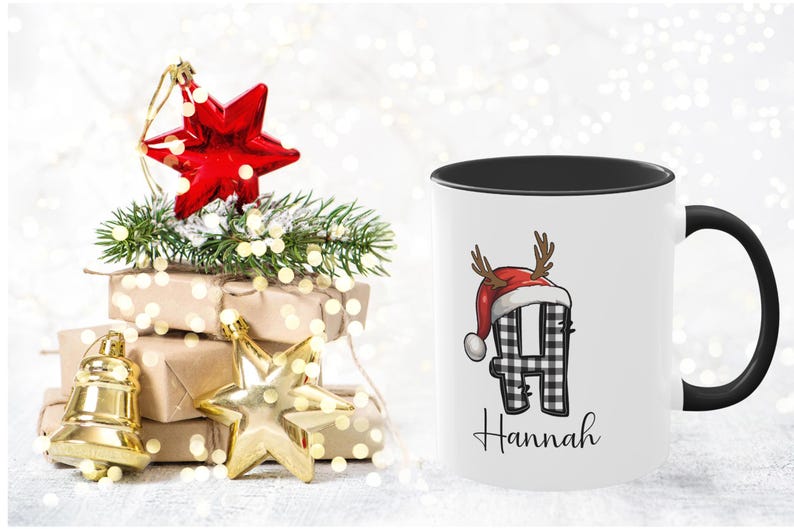 May include: White ceramic mug with a black interior and handle, featuring a plaid letter "H" wearing a Santa hat and antlers. The name "Hannah" is written below. Christmas decorations, including a red star ornament, are in the background.