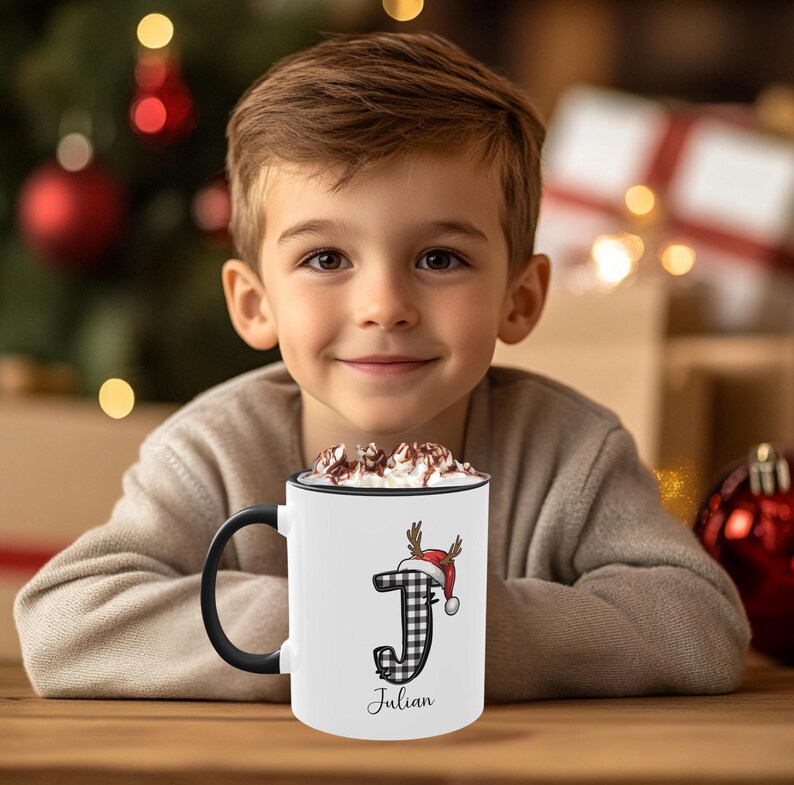 May include: White ceramic mug with a black handle and rim, featuring a black and white plaid letter "J" adorned with a reindeer antler and Santa hat design. The name "Julian" is printed below the letter. The mug is filled with a hot beverage.