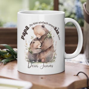 Op de afbeelding: Witte keramische mok met een aquarel illustratie van twee beren die elkaar omhelzen. De mok heeft Duitse tekst: "papa, Du bist einfach unentB&Auml;Rlich! Dein Jonas."