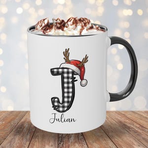 May include: White ceramic mug with a black handle and rim, filled with whipped cream and chocolate shavings. The mug features a black and white plaid letter "J" wearing a Santa hat with antlers, and the name "Julian" below.