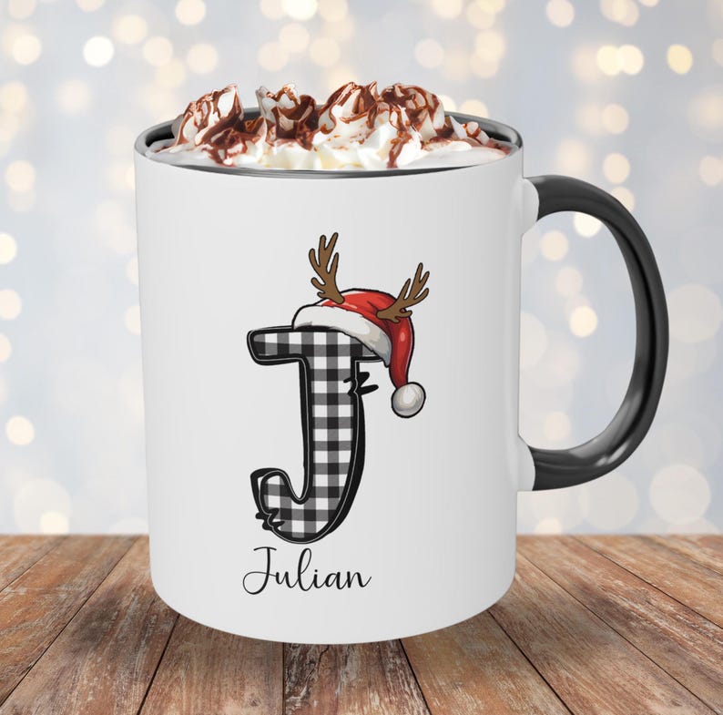 May include: White ceramic mug with a black handle and rim, filled with whipped cream and chocolate shavings. The mug features a black and white plaid letter "J" wearing a Santa hat with antlers, and the name "Julian" below.