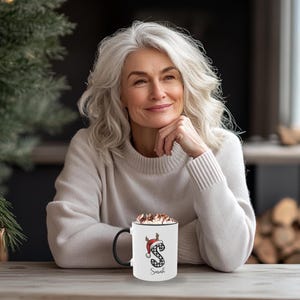 May include: A white coffee mug with a black handle, featuring a Christmas-themed design. The design includes a red Santa hat, antlers, and the letter "S" in a black and white plaid pattern. The name "Sarah" is printed below the letter.