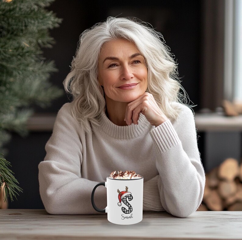 May include: A white coffee mug with a black handle, featuring a Christmas-themed design. The design includes a red Santa hat, antlers, and the letter "S" in a black and white plaid pattern. The name "Sarah" is printed below the letter.