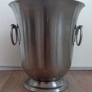 Può includere: Un secchiello per il ghiaccio in metallo argentato con due manici. Il secchiello ha un fondo arrotondato e una parte superiore larga e svasata.
