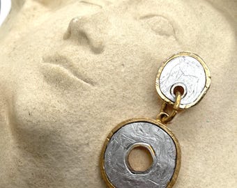 Puede incluir: Un pendiente colgante que presenta dos componentes circulares con un centro plateado texturizado y un borde dorado martillado. El círculo superior es sólido, mientras que el círculo inferior más grande tiene un recorte central.