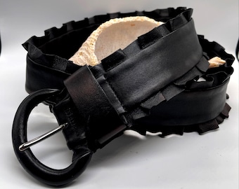 Puede incluir: Un cinturón de cuero negro con una hebilla grande y redonda y un borde con volantes. El cinturón está hecho de cuero grueso y resistente y tiene una hebilla de color plateado. El borde con volantes añade un elemento de diseño único.