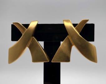 Puede incluir: Un par de pendientes dorados con un acabado mate cepillado. Cada pendiente tiene forma de letra X estilizada con dos bandas cruzadas, curvas y cónicas que crean una forma geométrica con bordes suaves, limpios y un aspecto sólido.