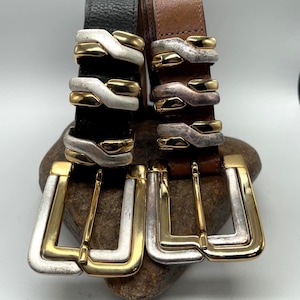 May include: Two leather belts with ornate silver and gold-tone buckles. One belt is black, the other is brown. The buckles have a unique design with layered metal accents. The belts are displayed on a stone.