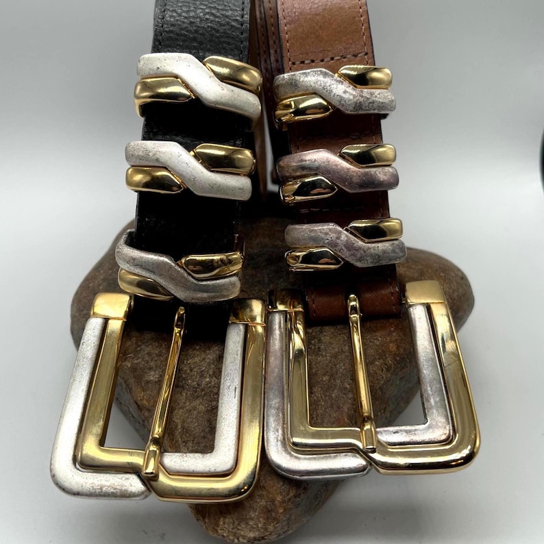 May include: Two leather belts with ornate silver and gold-tone buckles. One belt is black, the other is brown. The buckles have a unique design with layered metal accents. The belts are displayed on a stone.